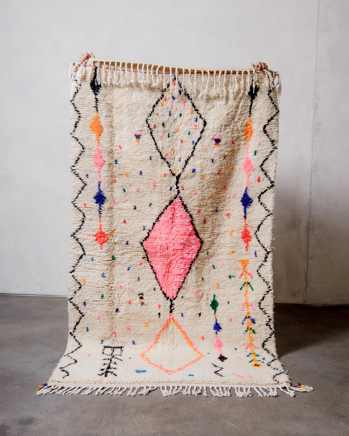 Berberlin Azilal rug made of fluffy cream wool with cotton tassels, held up against a white wall and cement floor.